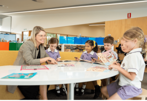 A photo of a teacher speaking to primary school students with books smiling in a very modern classroom facility